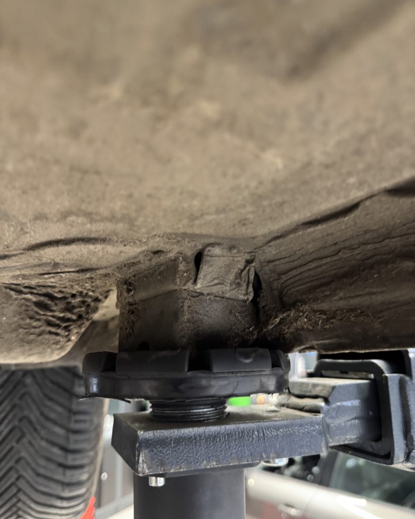 Ein schwarzer Hebebühnenstützpunkt hebt die Unterseite eines schmutzigen Fahrzeugs an, wobei sich ein Gummiklotz zwischen dem Stützpunkt und dem Fahrzeugrahmen befindet.
